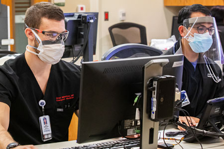 Two men in black scrubs and wearing PPE work at computers