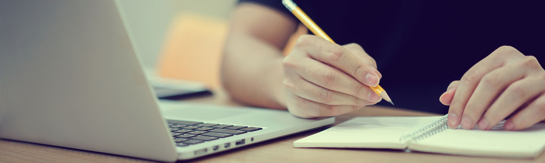 A woman writes with a pencil on a notebook beside a laptop