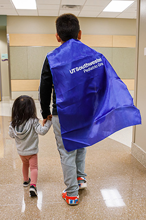 image of 2 children holding hands walkingi in hospital hallway, the taller child is wearing a superhero cape.