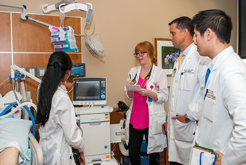 Four doctors at a patient's bedside. One woman points at a monitor as another takes notes, while two men at the right watch
