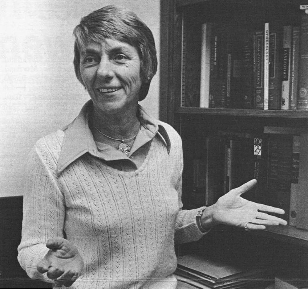 Black and white archive photo of Dr. Peggy Joyce Whalley standing in front of a bookcase with her hands turned upward