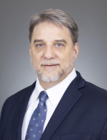 Man with graying hair, mustache and beard, wearing a dark blue suit, white shirt, and tie.