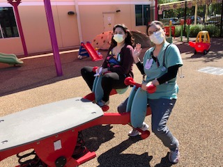 Two women wear masks while they sit on playground equipment