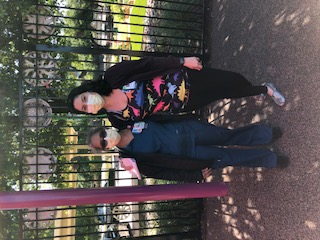 Two women wear masks and sit on a bench in front of an iron fence