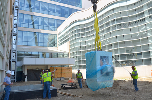 Construction workers guide heavy machine being carried by crane