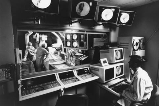 A man watches angiography images on monitors while procedure goes on in adjacent surgical room