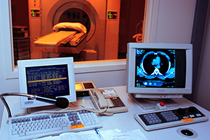 CT suite seen through window; desk in foreground shows scans on computer monitors