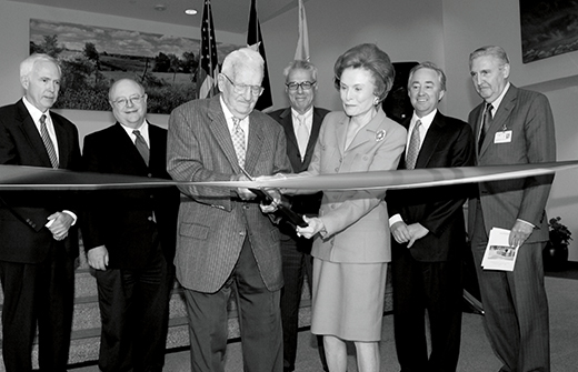 Bill Clements cuts ribbon with ceremonial scissors as Rita Clements assists and dignitaries look on