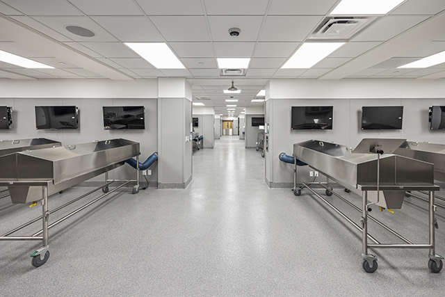 Wide view showing depth of the anatomy lab containing multiple rooms and learning stations with stainless steel tables and computer monitors.