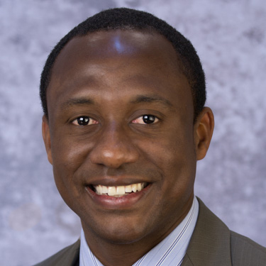 Meshka Anderson, smiling man with dark hair, wearing a suit and tie, close-up.