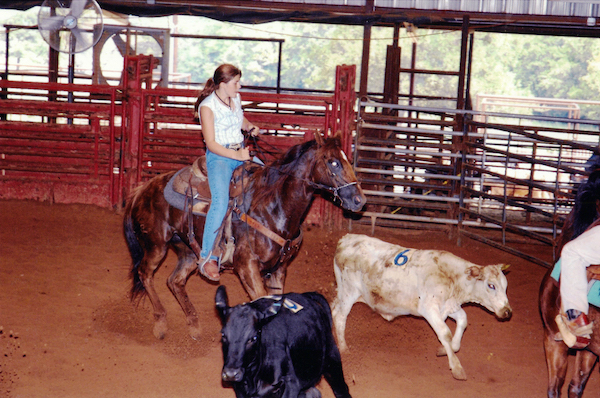 Chelsea rides a horse and rounds up cattle in a barn.