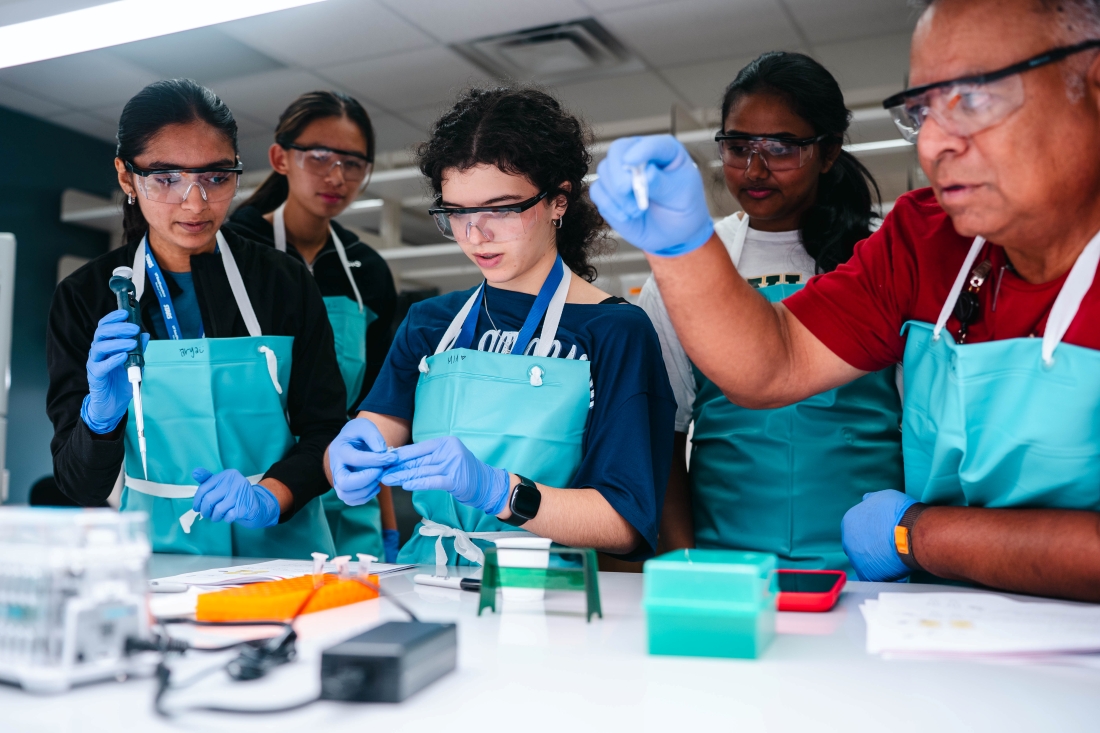 4 students and male instructor far right do experiments at a table while wearing safety glasses and aprons