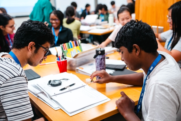 classroom of students at tables with 2 dark haired boys looking over notebook together