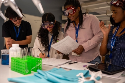 4 younger students in safety glasses share notes at a table with test tubes in a green rack