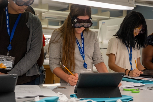 3 girls take notes at table while wearing safety glasses