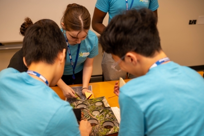 4 young students in blue tshirts work on craft project around a table