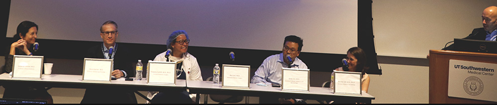 Five person panel seated at long table with one person standing at podium to right of table