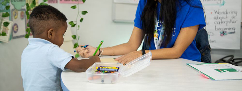 Young male student coloring with teacher at table in classroom