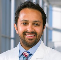 Smiling dark-haired man with beard and mustache wearing a lab coat.