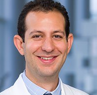 Smiling dark-haired man wearing a white lab coat.