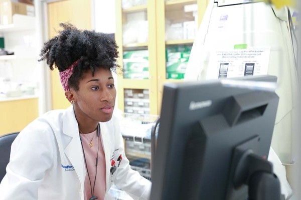 young woman wearing white lab coat sits at computer keyboard and monitor
