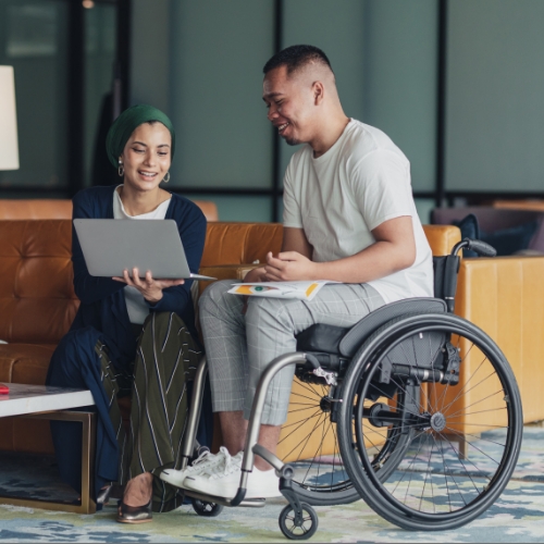 A female holding a laptop, smiling, talking to a man in a wheelchair