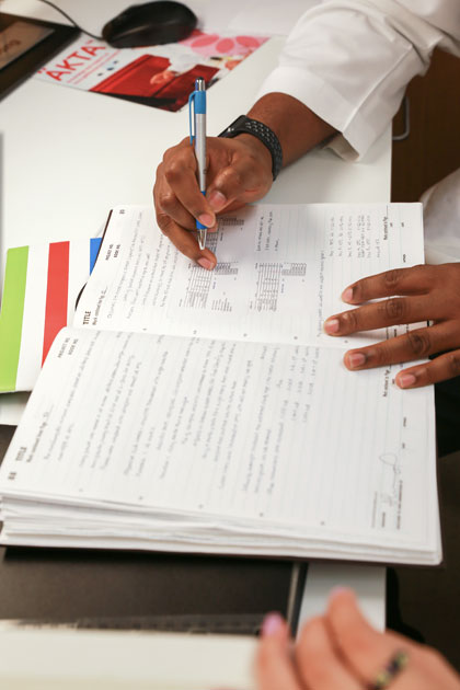 One hand holding an ink pen as two people look over a notebook