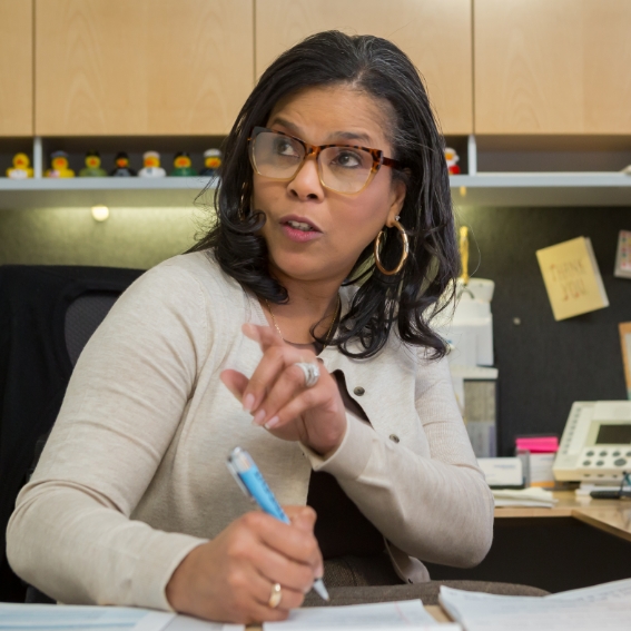 Close up of female sitting at desk with a pen in her right hand. She is looking at and speaking with someone outside of the camera frame.