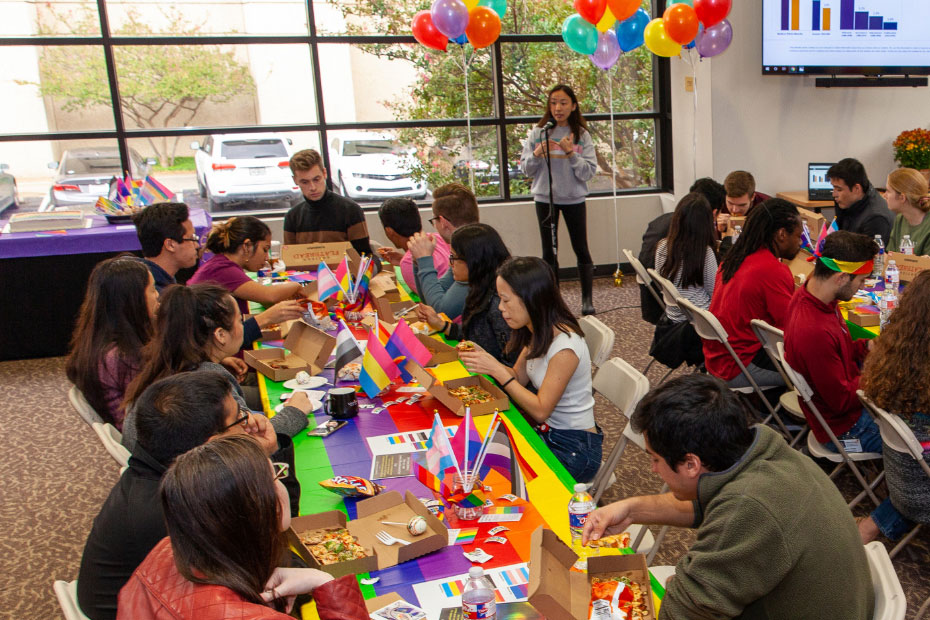 A woman speaks at a microphone as students eat lunch with rainbow table cloths on the tables