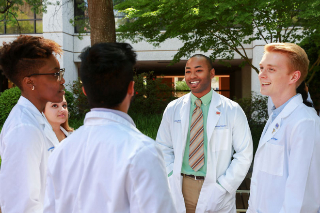 Three male and two female medical students in white coats gather outside