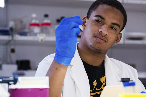 A Black male in a white coat holds a test tube with a gloved hand