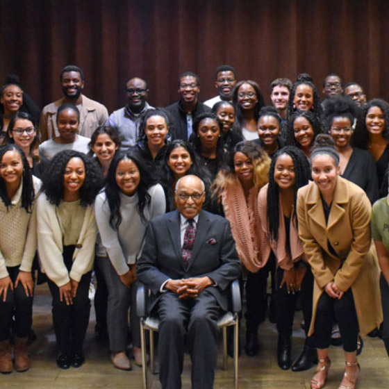 A group of Black people stand around a Black man who is sitting in a chair