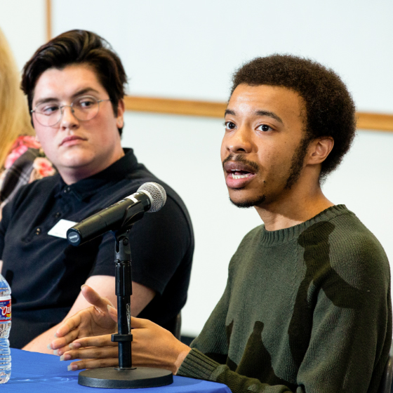 A Black man speaks at a microphone as a white man watches from beside him