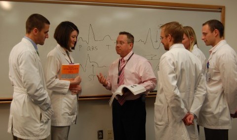 Students in white coats listen as a male faculty member makes a point
