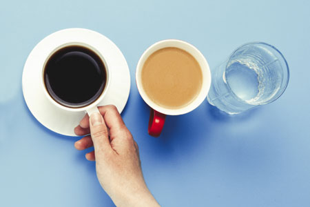 A woman's hand reaching for a cup of coffee that's sitting on a saucer with a cup of tea and a glass of water beside it
