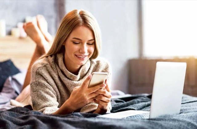 girl reclines on bed while working on cell phone and laptop