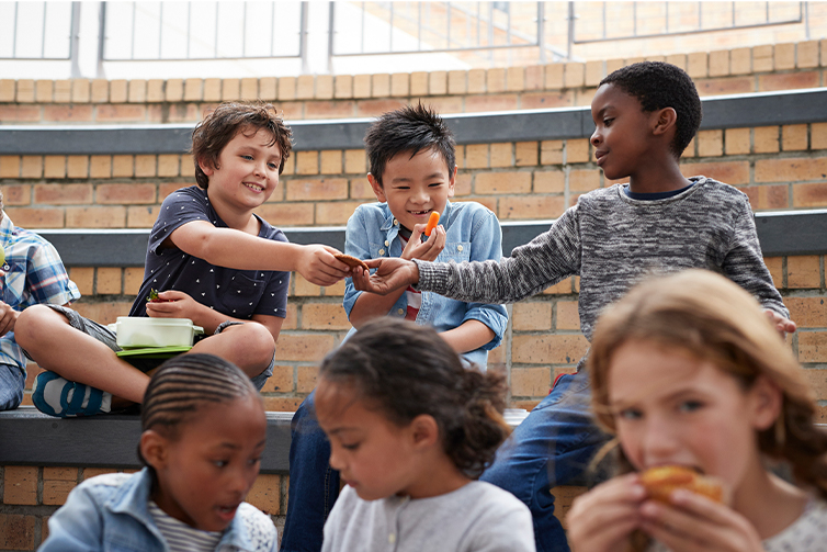 Elementary school aged boys and girls eat lunch with one boy handing another a cookie and a boy giggles as he eats a carrot