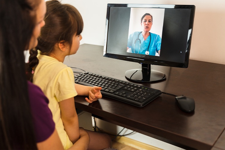A woman with a young child on her lap speaks with a female doctor on a computer monitor