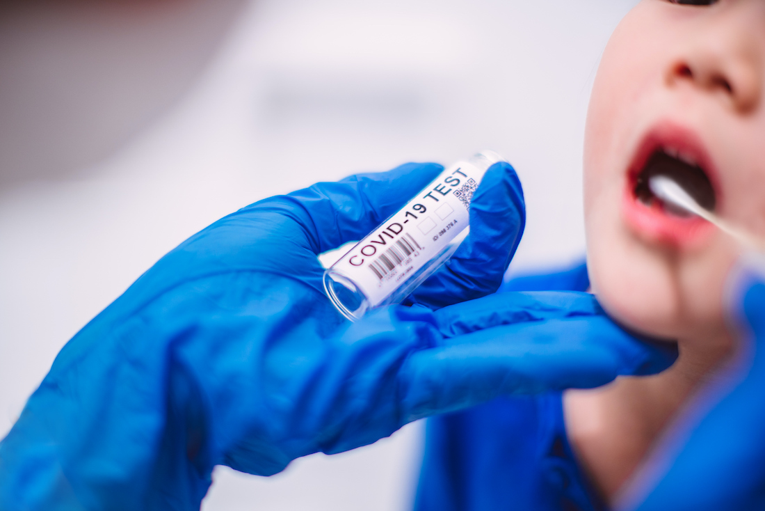 Doctor’s hands in protection gloves putting COVID-19 test swab into kid’s mouth in hospital.