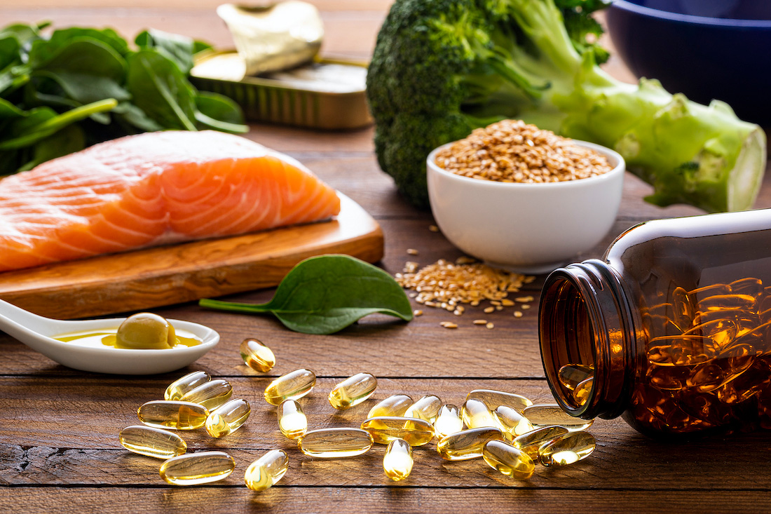 Front view of many fish oil capsules spilling out from the bottle surrounded by an assortment of food rich in omega-3 such as salmon, flax seeds, broccoli, sardines, spinach, olives and olive oil.