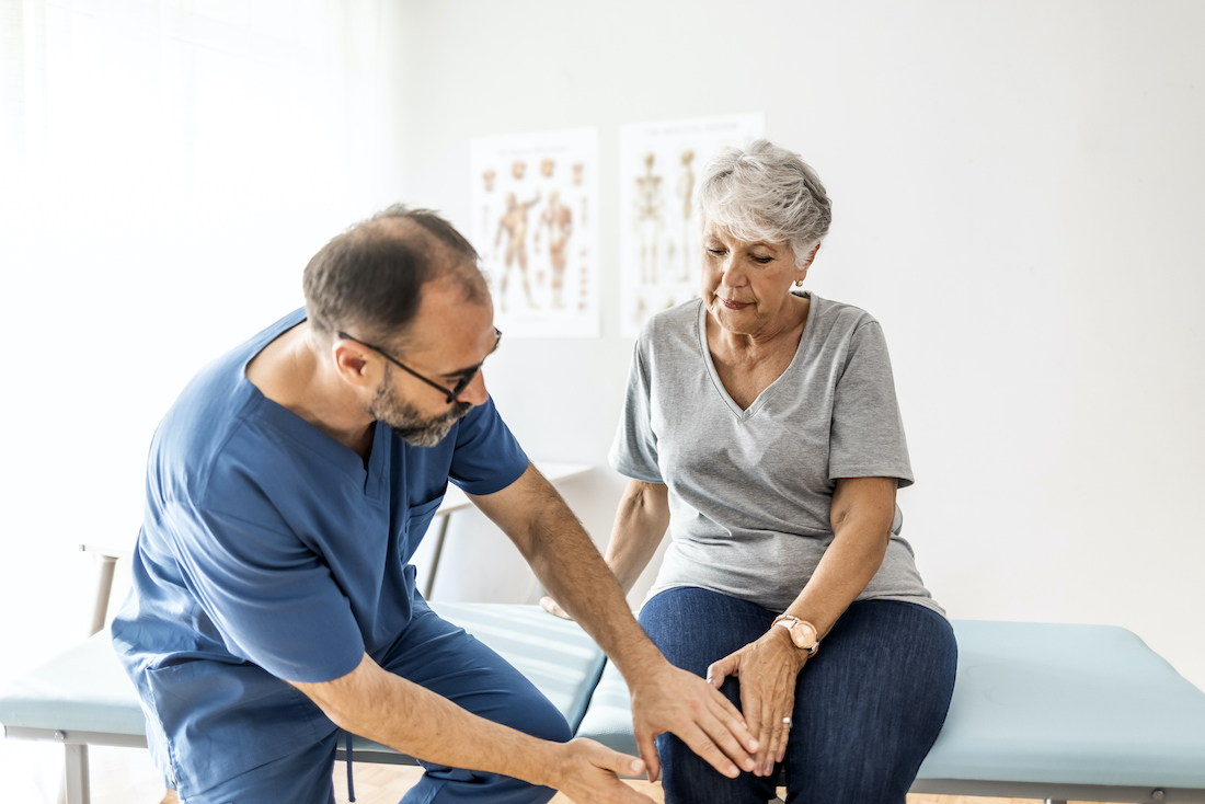 Photo of senior woman having some knee pain. She's at doctor's office having medical examination by a male doctor. The doctor is touching the sensitive area and trying to determine the cause of pain.