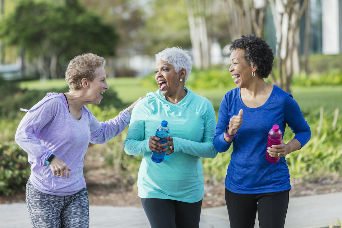 A group of three women exercising in the park, talking and laughing as they power walk, carrying water bottles.