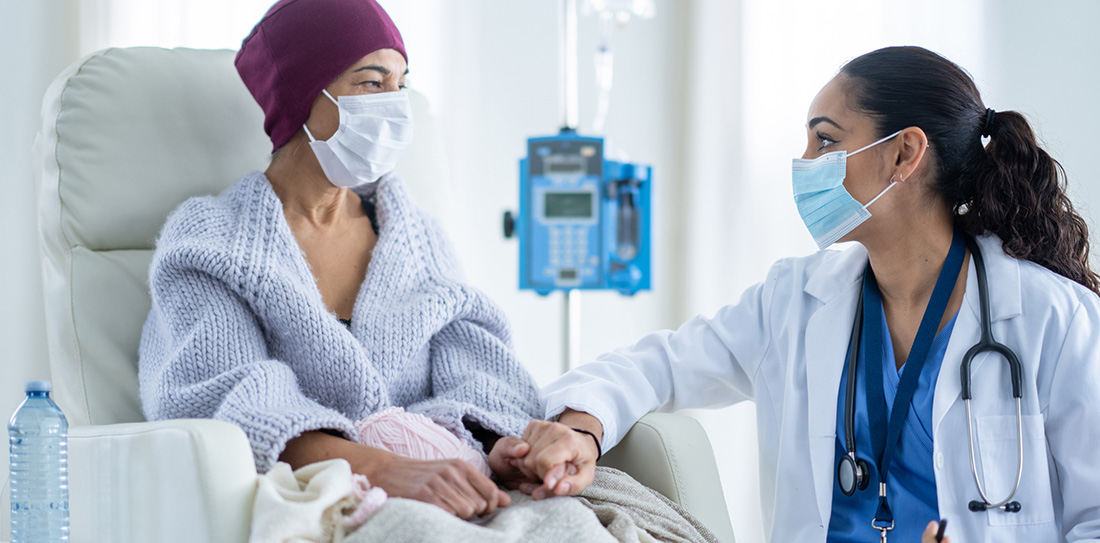 Female cancer patient sitting in chair in chemo treatment room looking at female doctor who is holding her hand