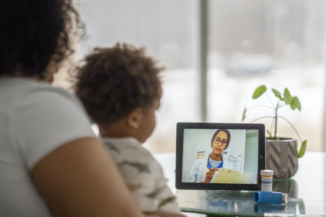 African American mother sitting with her sick child and speaking with a medical professional via video chat during the COVID-19 pandemic.