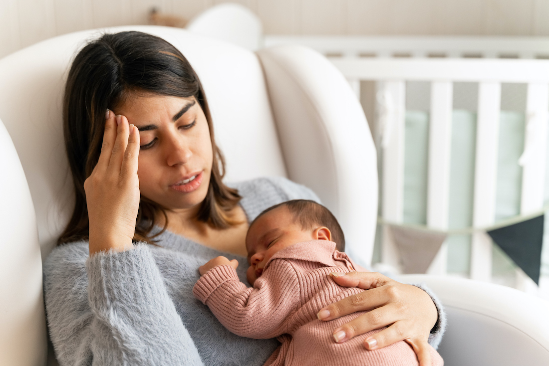 Mother sitting on couch in nursery feeling postpartum depression with baby in her arms.