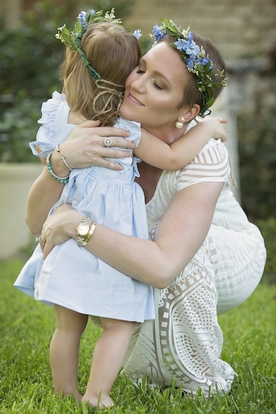 Ann Marie Herbst hugs her daughter, Hayden. They are outdoors; both are wearing summery dresses with flower headdresses.