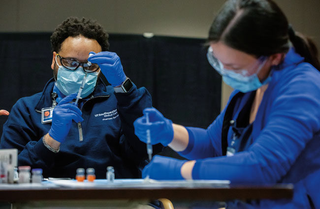 Two UTSW nurses in blue scrubs, blue masks, gloves, and glasses fill syringes of COVID-19 vaccines from vials