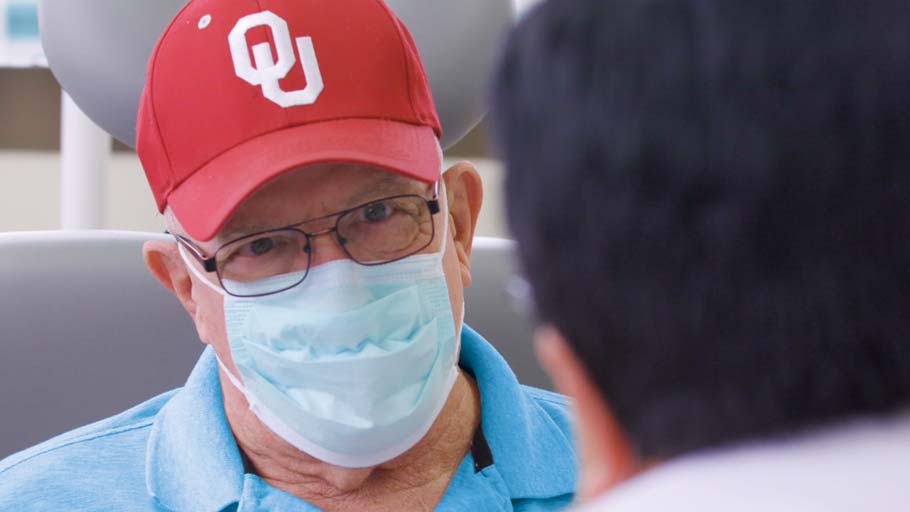 A man wears a blue face mask and a red University of Oklahoma hat as he talks with a doctor