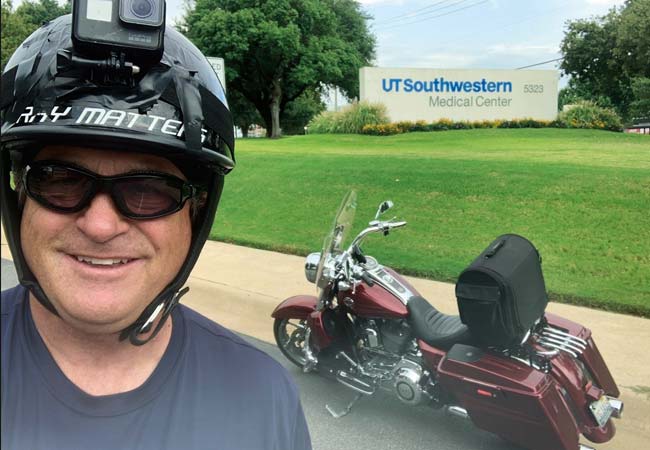A man wearing a black motorcycle helmet and glasses stands in front of his motorcycle on the UT Southwestern campus