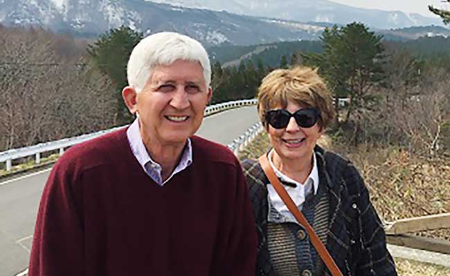 Kent and JoAnn “Jodi” Foster pose in front of a roadway with a beautiful mountain backdrop